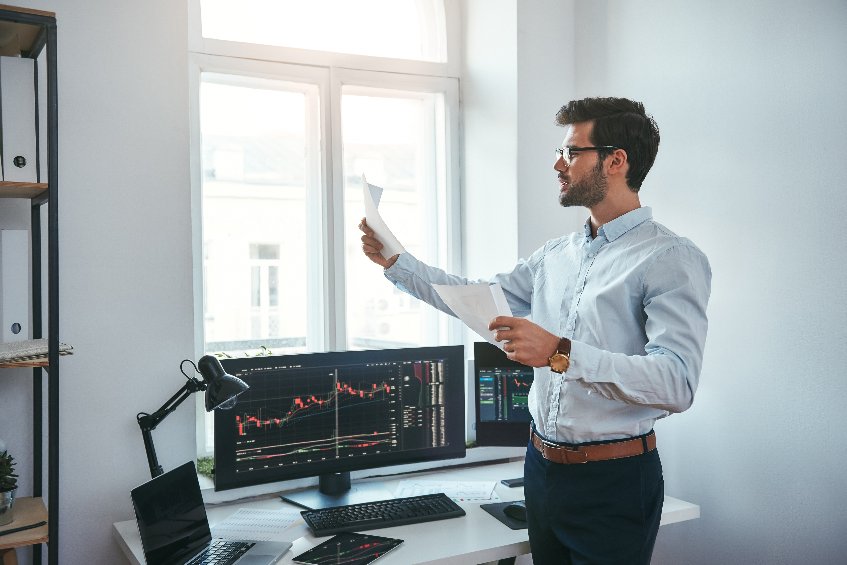 young trader in eyeglasses looking at financial reports and analyzing trading charts while standing in front of computer screens in office