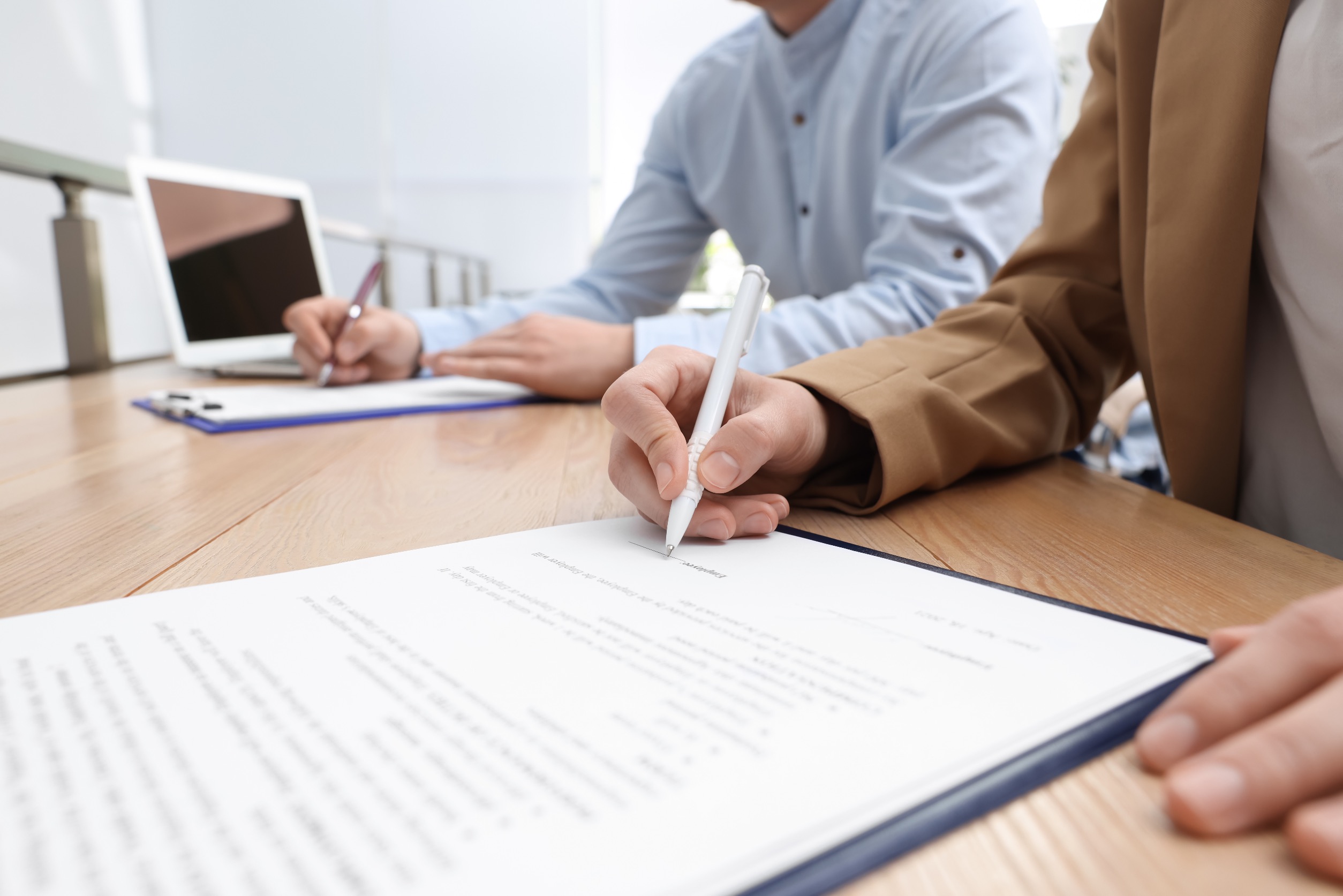 Two people signing documents on clipboards in a professional setting - the foreground subject is wearing a khaki blazer and white shirt while the background subject wears a button down blue shirt and is next to an open white laptop.